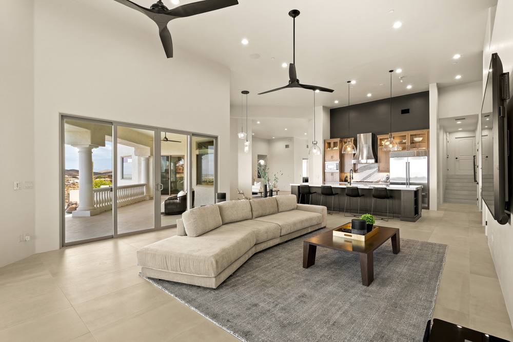 A bright, airy living room with a large L-shaped beige sofa, wooden coffee table, and floor-to-ceiling glass doors that open to a covered patio. The space flows into a modern kitchen with dark cabinetry and pendant lights, creating a seamless indoor-outdoor experience.